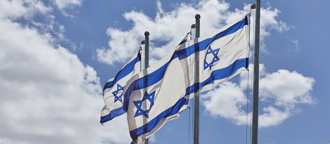 Four Israeli flags waving under the blue, cloudy sky with hills. Blue and white flags with Star of David symbolize Israels independence. Low angle view, flags flying high, a patriotic symbol.
